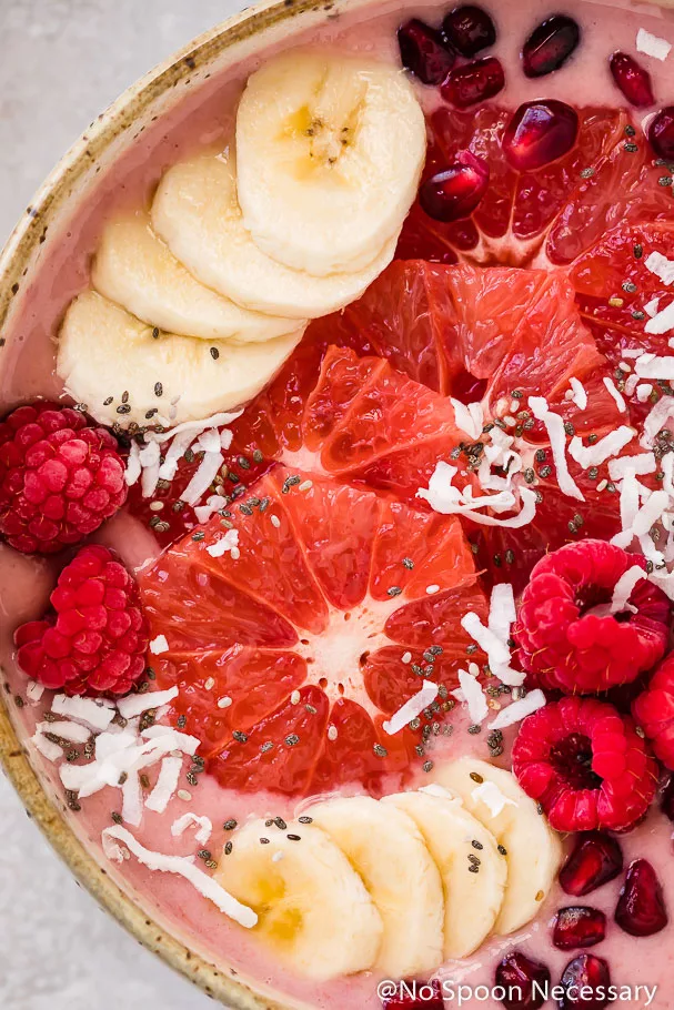 Overhead, extreme up-close shot of a Berry Citrus Smoothie Bowl topped with slices of bananas, grapefruit, whole raspberries, pomegranate arils, chia seeds and shredded coconut.