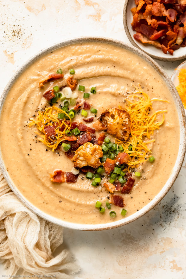 Overhead photo of a bowl of creamy roasted cauliflower soup topped with cheese, bacon and sliced scallions.