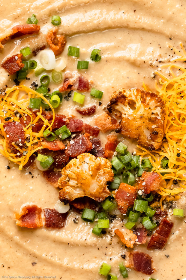 Overhead, close-up photo of creamy cauliflower soup topped with roasted cauliflower florets, crispy bacon, shredded cheese and sliced scallions.