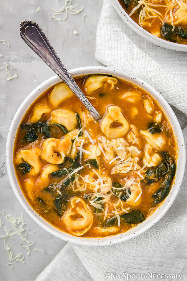 Overhead shot of a bowl of Chicken, Spinach and Tortellini Tomato Soup topped with shredded parmesan with a spoon inserted into the bowl and a light silver linen and another bowl of soup off to the side.