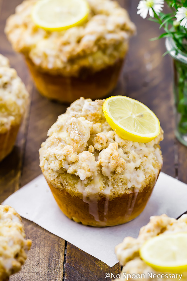 Angled, straight on shot of a Jumbo Lemon Crumb Glazed Muffin on a square piece of parchment paper with more muffins and a small jar of field flowers surrounding the muffin.