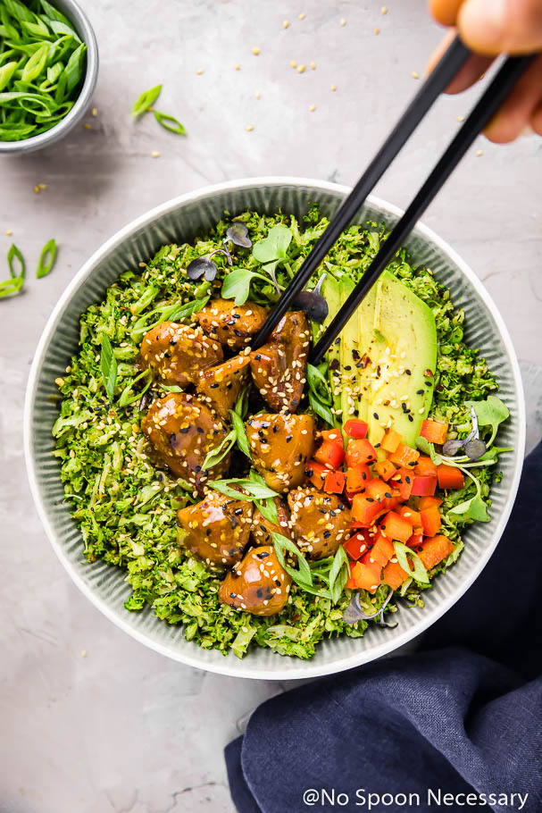 Overhead shot of a hand holding black chop sticks picking up a piece of Slow Cooker Honey Sesame Chicken from a bowl filled with broccoli rice, honey sesame chicken, sliced avocado and diced red pepper with a blue linen and a ramekin of sliced scallions framing the shot.