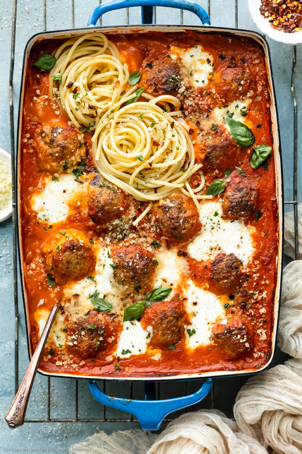 Overhead photo of spicy chicken meatballs baked in vodka tomato sauce and topped with cheese and cooked spaghetti in a large 9x13-inch pan with a large serving spoon inserted into the pan of meatballs.
