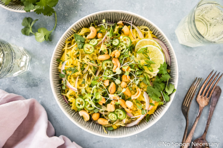 Overhead, landscape shot of a bowl of healthy Spicy Thai Spaghetti Squash Salad garnished with slices of red onion, jalapenos, crushed cashews, cilantro and lime slices; with a pale purple linen, forks, wine glasses, small bowl of cilantro and lime wedges surrounding the bowl.