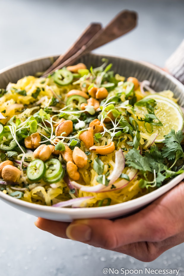 45 degree angle shot of two hands holding a bowl of Spicy Thai Spaghetti Squash Salad garnished with slices of red onion, jalapenos, crushed cashews, cilantro and lime slices; with a fork and spoon tucked into the back of the bowl.
