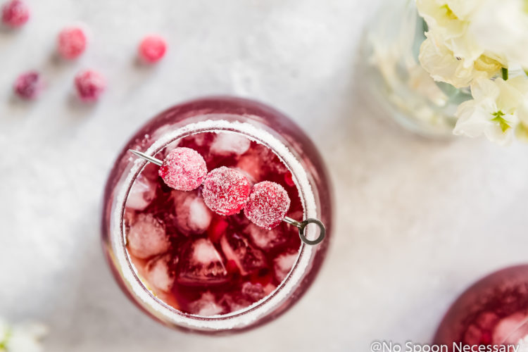 Overhead shot of a cocktail glass filled with Cranberry Pomegranate Margarita with a salt & sugar rim and garnished with sugared cranberries; with a small vase of white flowers, sugared cranberries and an additional cocktail surrounding the glass.