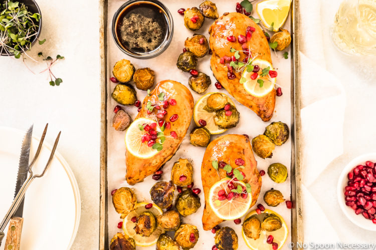 Overhead shot of a Sheet Pan containing Honey Balsamic Chicken & Brussels Sprouts garnished with lemon slices, pomegranate arils and micro greens; with a stack of plates, knife, meat fork, glass of wine, and small bowls of pomegranate arils and micro greens surrounding the pan.
