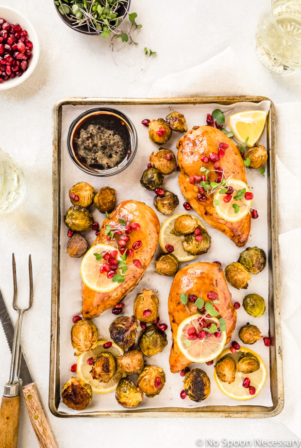Overhead shot of a Sheet Pan containing Honey Balsamic Chicken & Brussels Sprouts garnished with lemon slices, pomegranate arils and micro greens; with a knife, meat fork, glasses of wine, and small bowls of pomegranate arils and micro greens surrounding the pan.