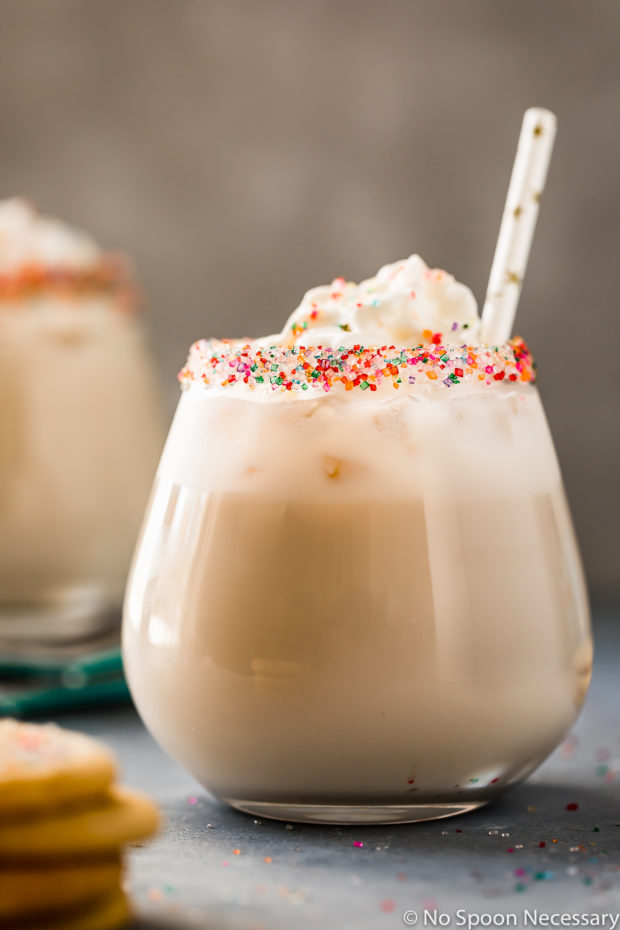Straight on shot of a Sugar Cookie Cocktail topped with whipped cream and a straw in a sprinkle rimmed glass with a small stack of cookies and an additional cocktail barely visible and blurred around the glass.