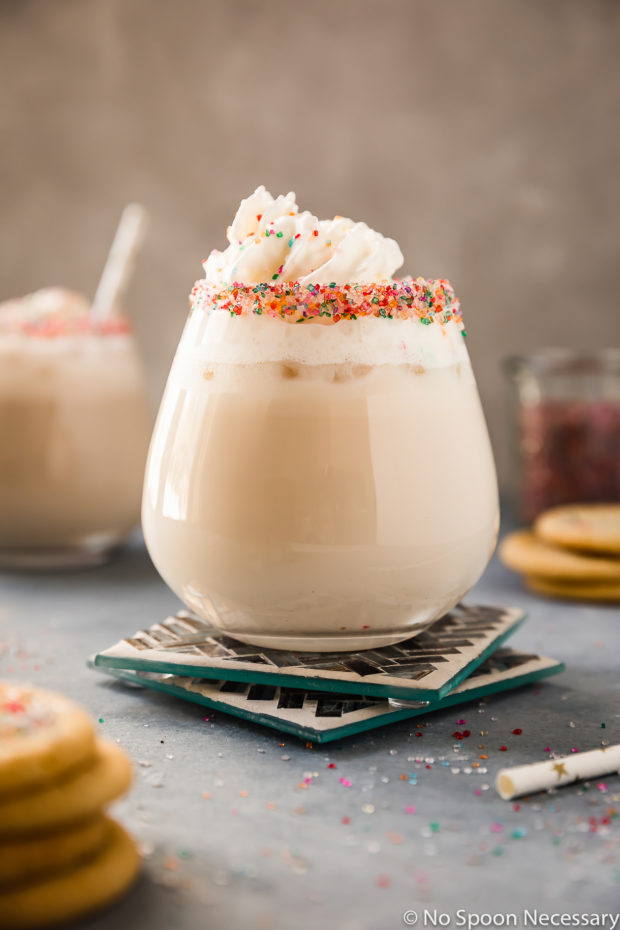 Straight on shot of a Sugar Cookie Cocktail topped with whipped cream in a sprinkle rimmed cocktail glass on 2 blue tiled coasters with miniature sugar cookies, a straw, an additional cocktail and a jar of sprinkles surrounding the glass.
