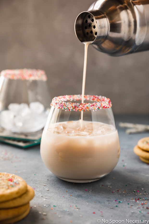 Straight on shot of a Sugar Cookie Cocktail being poured from a metal shaker into a sprinkle rimmed cocktail glass with miniature sugar cookies, sprinkles, straws and an additional cocktail glass filled with ice surrounding the glass.