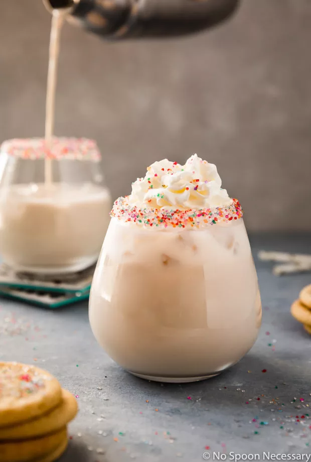 Straight on shot of a Sugar Cookie Cocktail topped with whipped cream in a sprinkle rimmed cocktail glass with miniature sugar cookies, a straw, an additional glass being filled with a sugar cookie cocktail from a metal shaker blurred around the glass.