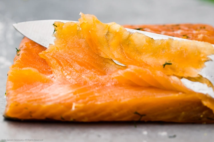 Straight on, landscape photo of a filet of salmon gravlax with a slicing knife and one single, thinly sliced piece of gravlax laying on top of the filet.