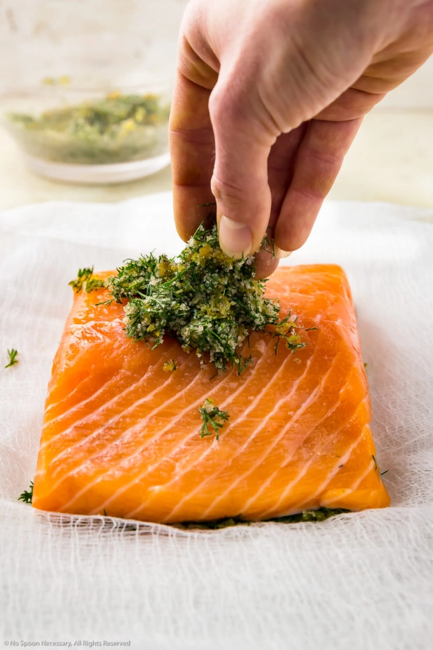 Straight on photo of a hand putting a dry cure of citrus zest and dill on top of a filet of salmon - step 4 of the recipe.