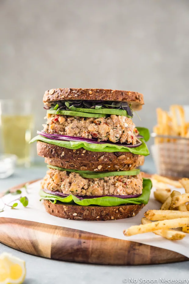 Straight on shot of two Mediterranean Smashed Chickpea Salad Sandwiches with micro greens, sliced avocado, red onions and lettuce on whole grain bread stacked on top of each other on a wood platter with a basket of french fries and glass of beer blurred in the background.