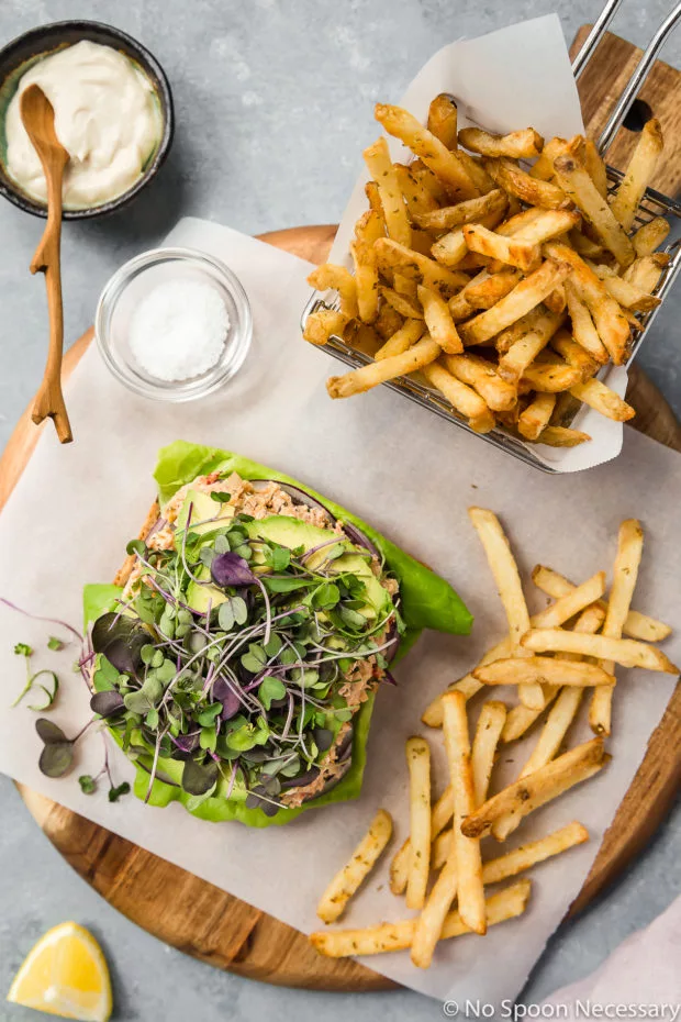 Overhead shot of an open faced Mediterranean Smashed Chickpea Salad Sandwich with micro greens, sliced avocado, red onions and lettuce a wood platter lined with parchment paper with a basket of french fries, ramekin of tahini mayonnaise, pinch bowl of salt and lemon wedge surrounding the sandwich. 