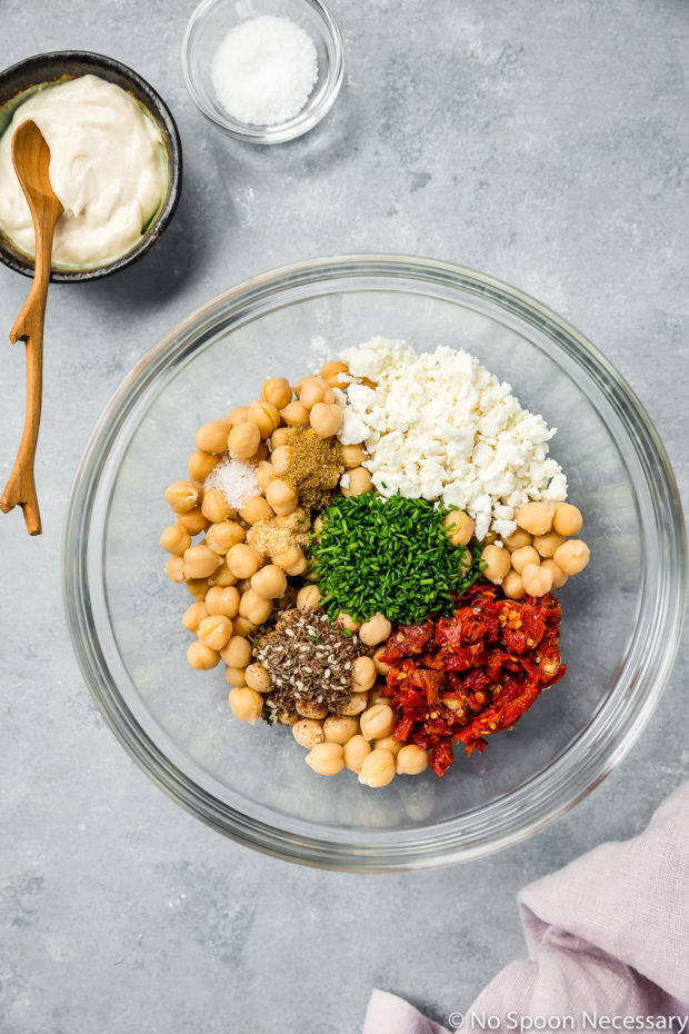 Overhead shot of all the ingredients needed to make Mediterranean Smashed Chickpea Salad in a glass bowl with a ramekin of tahini mayonnaise, pinch bowl of salt and purple linen surrounding the bowl - 1st step in the recipe of how to make a Mediterranean Smashed Chickpea Salad Sandwich.