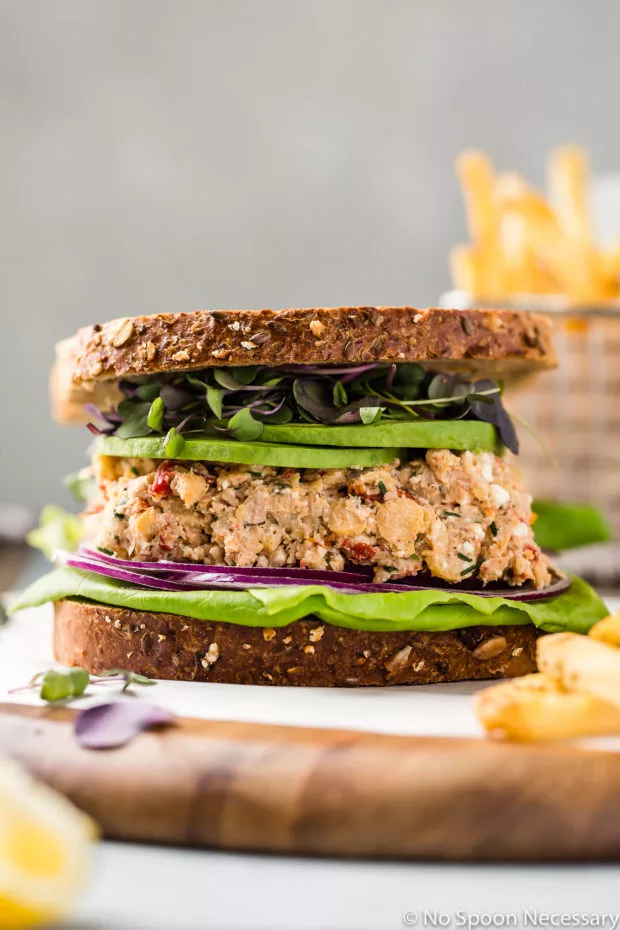 Straight on shot of a Mediterranean Smashed Chickpea Salad Sandwich with micro greens, sliced avocado, red onions and lettuce with whole grain bread on a wood platter with a basket of french fries blurred in the background.