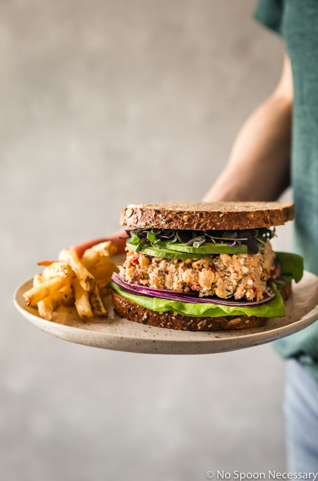 Straight on shot of a person holding a plate containing a Mediterranean Smashed Chickpea Salad Sandwich with micro greens, sliced avocado, red onions and lettuce on whole grain bread with french fries off to the side of the sandwich.
