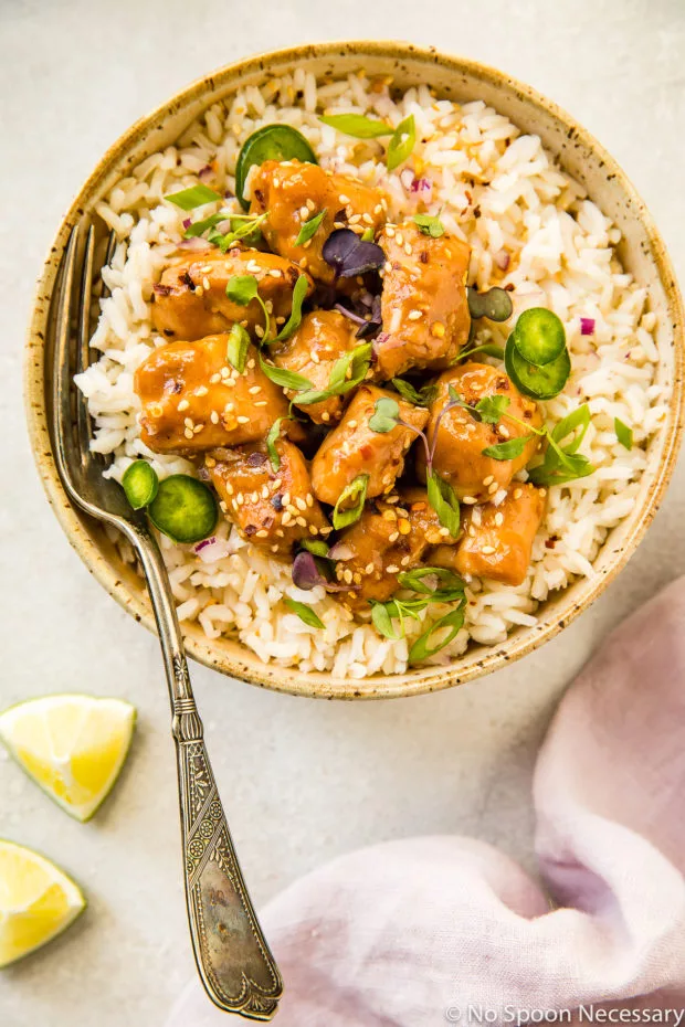 Overhead shot of a neutral colored bowl filled with rice and topped with Slow Cooker Bourbon Plum Chicken, with a fork inserted into the rice and a pale purple linen and lime wedges tucked in the bottom corners of the shot.