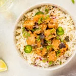 Overhead, landscape shot of a white bowl filled with rice and topped with Slow Cooker Bourbon Plum Chicken, with a pale purple linen, lime wedges, small bowl of sesame seeds, wine glass and sliced scallions surrounding the bowl.