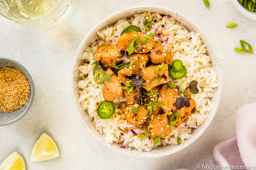 Overhead, landscape shot of a white bowl filled with rice and topped with Slow Cooker Bourbon Plum Chicken, with a pale purple linen, lime wedges, small bowl of sesame seeds, wine glass and sliced scallions surrounding the bowl.