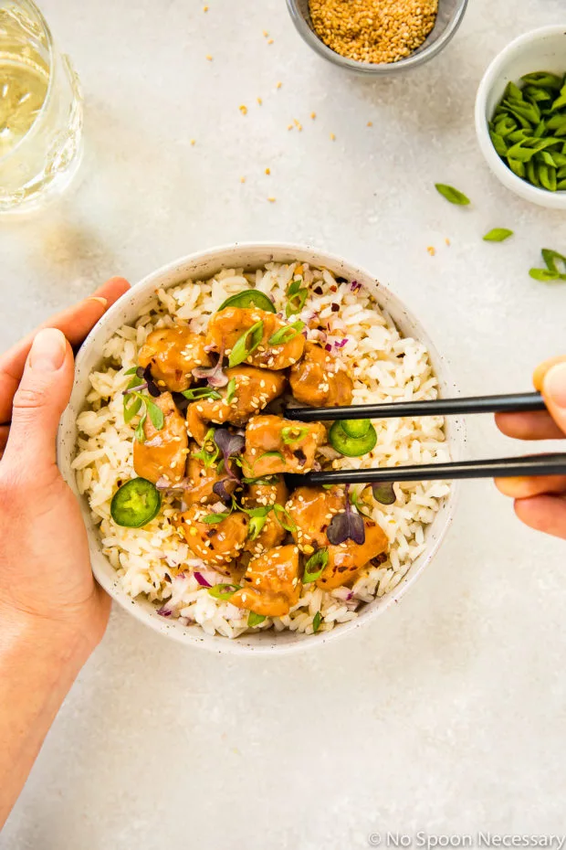 Overhead shot of a neutral colored bowl filled with rice and topped with Slow Cooker Bourbon Plum Chicken, with a hand holding the bowl and another hand holding chopsticks picking up a piece of chicken, with a glass of wine and small bowls of sesame seeds and sliced scallions tucked into the upper corners of the shot.