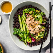 Overhead, landscape shot of a neutral colored serving bowl filled with Asian Avocado Tuna Salad Grain Bowl with black chopsticks resting on the top of the bowl and a purple linen, small additional salad, ramekin of honey miso dressing and ramekin of sesame seeds surrounding the serving bowl.