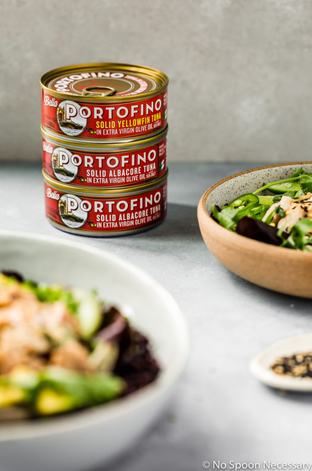 Angled shot of a stack of three cans of Bella Portofino Tuna with two Asian Avocado Tuna Salad Grain Bowls blurred in front of the cans.