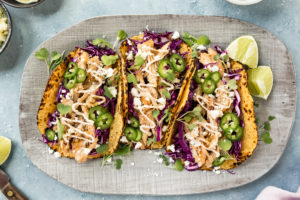 Overhead, landscape photo of a gray platter of Beer Chicken Tacos on a blue surface with a pale purple linen, lime wedges, small knife, and bowls of salt, cotija cheese and baja sauce surrounding the platter.