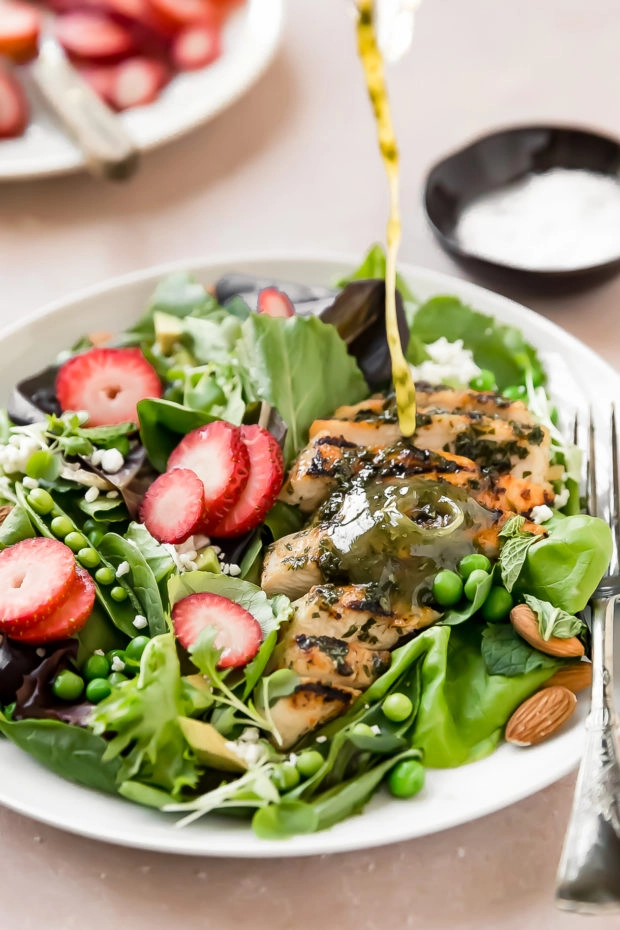 Angled, up-close shot of a herb vinaigrette being drizzled on a Strawberry Salad with Grilled Chicken on white plate with a fork; with a plate of sliced strawberries and small ramekin of salt blurred in the background.
