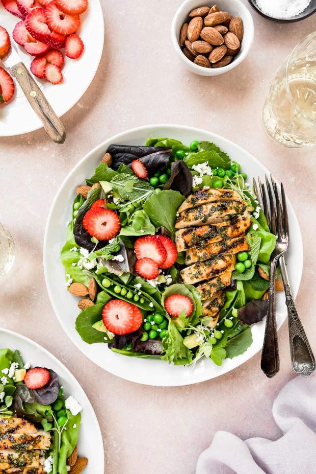 Overhead shot of two Strawberry Salads with spinach and grilled chicken on white plates with a plate of sliced strawberries, wine glasses, pale purple linen, bowl of almonds, and small ramekin of salt surrounding the plates.