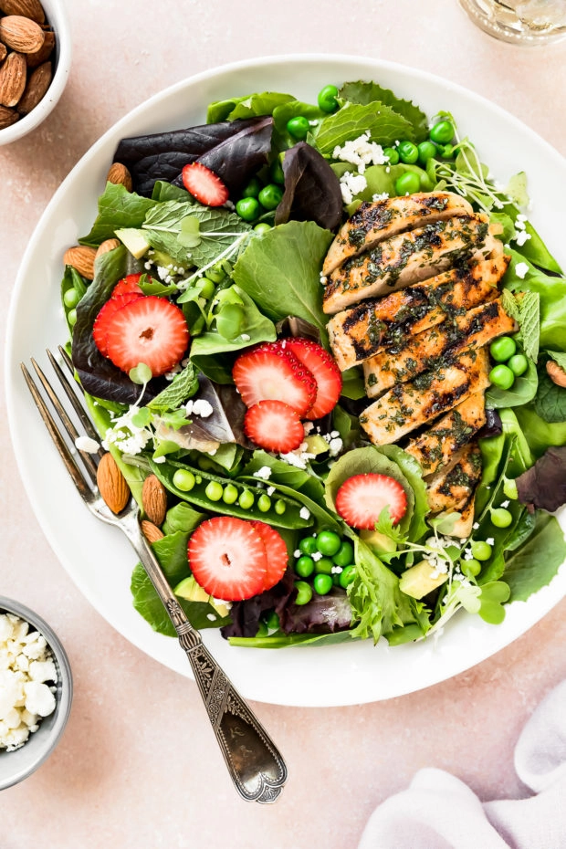 Overhead photo of a Fresh Strawberry Spinach Salad topped with grilled chicken on a white plate with a fork inserted into the salad and ramekins of crumbled goat cheese and almonds next to the plate.