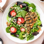 Overhead, landscape shot of a Strawberry Spinach Salad with Grilled Chicken on a white plate with a plate of sliced strawberries, wine glasses, pale purple linen, bowl of almonds, and small ramekin of salt, bowl of feta and forks surrounding the plates.