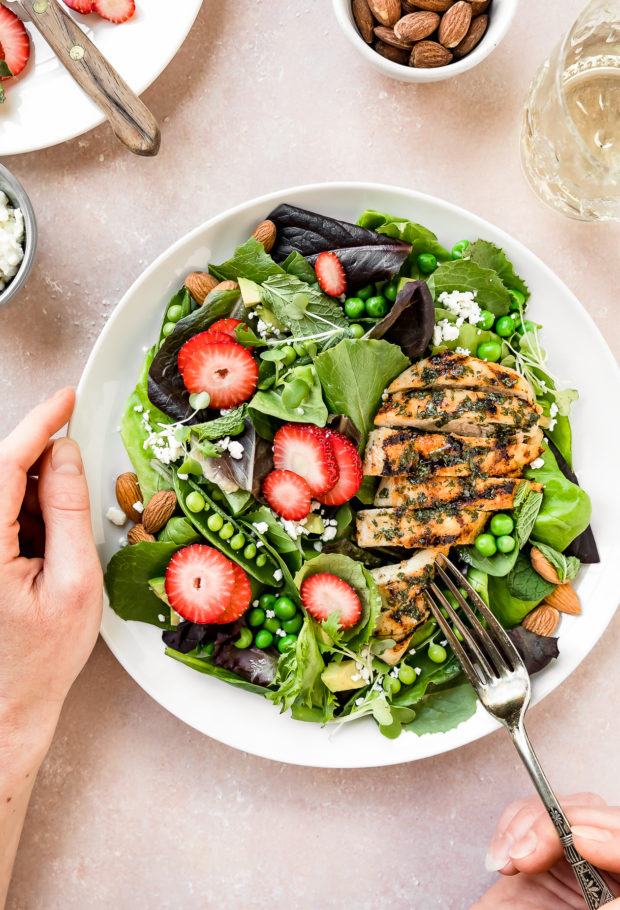 Overhead photo of Spinach Strawberry Salad with Grilled Chicken on white plate with one hand holding the plate and another hand holding a fork inserted into the chicken; with a plate of sliced strawberries, bowl of almonds, bowl of feta and wine glass surrounding the plate.
