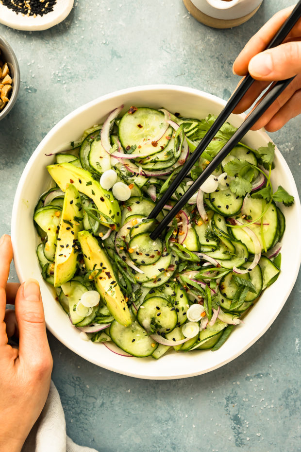 Overhead photo of Fresh Cucumber Salad in a white bowl with one hand holding the bowl and another hand holding black chopsticks picking up some of the salad.