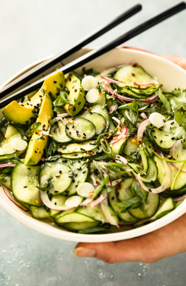 Angled photo of two hands holding a white bowl filled with Asian Cucumber Salad with sesame seeds, chopped cilantro and sliced scallions with a pair of black chopsticks resting on the side of the bowl.