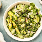 Overhead photo of Cucumber Salad in a white bowl with a jar of Asian dressing, neutral linen and ramekins of sesame seeds and peanuts surrounding the bowl.