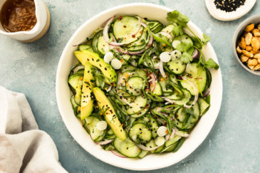 Overhead photo of Cucumber Salad in a white bowl with a jar of Asian dressing, neutral linen and ramekins of sesame seeds and peanuts surrounding the bowl.