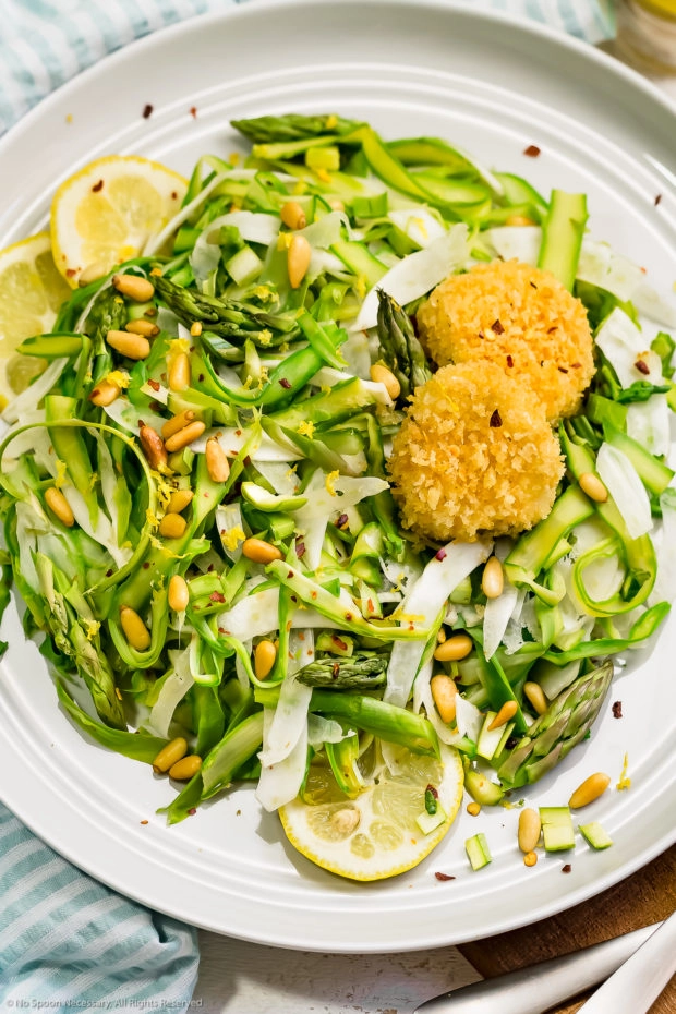 Overhead photo of Shaved Raw Asparagus Salad topped with baked goat cheese, pine nuts and lemon slices on a gray dinner plate.