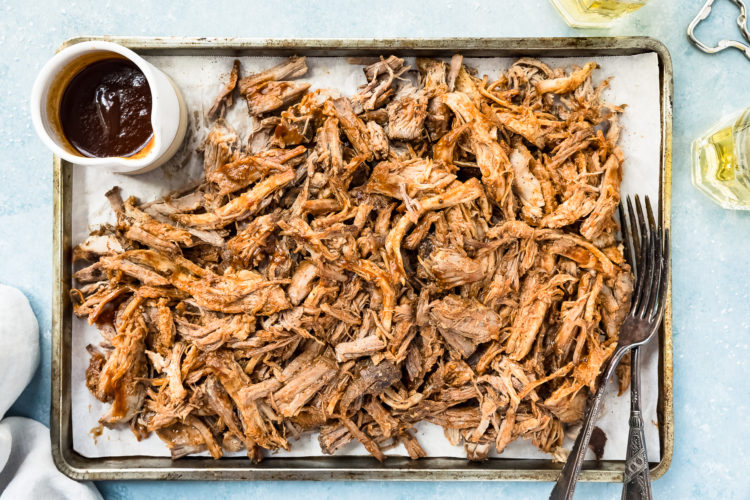 Overhead, landscape shot of Easy Carolina BBQ Pulled Pork on a small sheet pan with a ramekin of BBQ sauce and two forks; with two glasses of beer and a bottle cap opener off to the side of the pan.