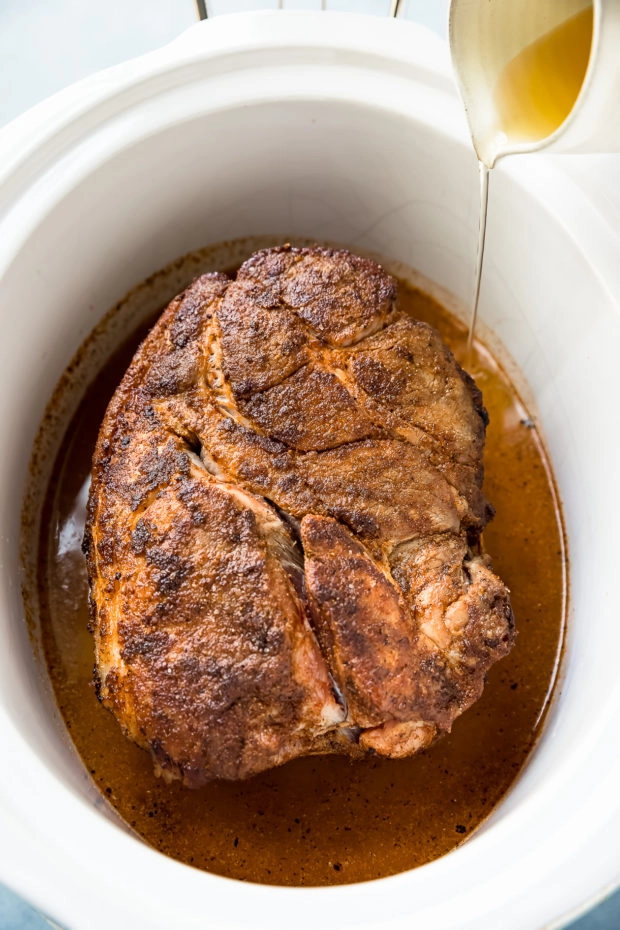 Angled shot of a seared pork butt in a slow cooker with a small container of apple cider being poured into the slow cooker. (A photo of step 3 in the recipe to make Slow Cooker Easy Carolina BBQ Pulled Pork)