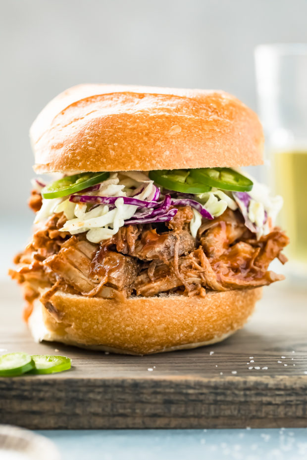 Straight on shot of Easy Carolina BBQ Pulled Pork sandwich on a brioche bun with Jalapeno Coleslaw; with a glass of beer blurred and barely visible in the background.
