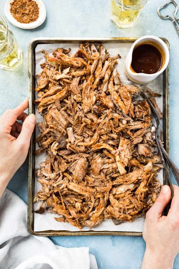 Overhead shot of Easy Carolina BBQ Pulled Pork on a small sheet pan with a ramekin of BBQ sauce and two forks; there are two hands holding the sheet pan and two glasses of beer, a bottle cap opener and a small ramekin of BBQ rub off to the side of the pan.