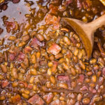 Overhead photo of easy baked beans made with bacon and brown sugar in a large white pot with a wooden spoon stirring the beans.