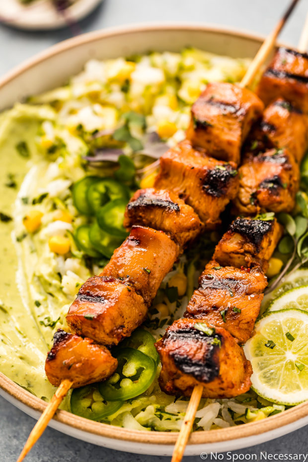 Angled, up close shot of Grilled Honey Chipotle Chicken Bowls consisting of two grilled chicken skewers, corn studded zucchini rice, avocado cilantro sauce and sliced limes; with the focus of the shot on the chicken skewers.