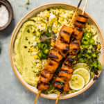 Overhead shot of Grilled Honey Chipotle Chicken Bowls consisting of two grilled chicken skewers, corn studded zucchini rice, avocado cilantro sauce and sliced limes; with a neutral linen, glass of white wine and ramekins of salt and microgreens surrounding the bowl.