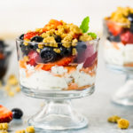 Straight on shot of two fruit Parfaits - which consists of layers of whipped Greek yogurt, freesh fruit and graham cracker streusel - in a small parfait glass, with an additional parfait, bowl of fruit and fresh fruit strewn around the parfait.