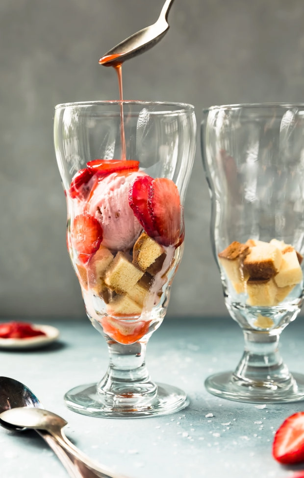 Straight on shot of a sundae glass filled with cubed pound cake, a scoop of strawberry ice cream and amaretto strawberries with a spoon drizzling amaretto sauce over the top; with additional spoons, another sundae glass and fresh strawberries surrounding the glass. (Photo of step 3 of the recipe)