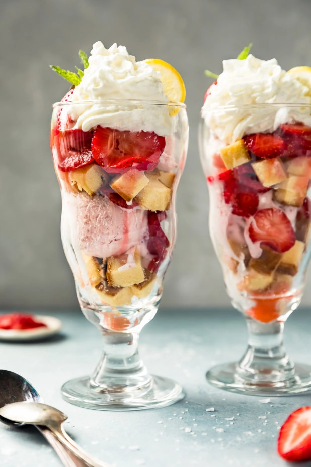 Straight on shot of two homemade Strawberry Shortcake Ice Cream Sundaes with spoons, coarse salt, and a small ramekin of sliced strawberries arranged next to the sundaes.